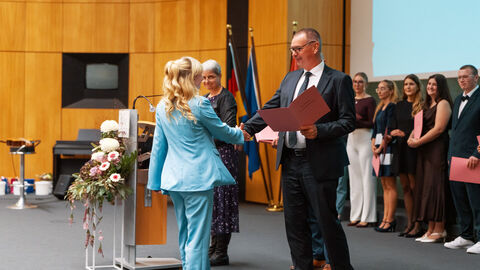 Ein Mann schüttelt auf einer Bühne einer Frau die Hand und überreicht ihr eine rosafarbene Mappe